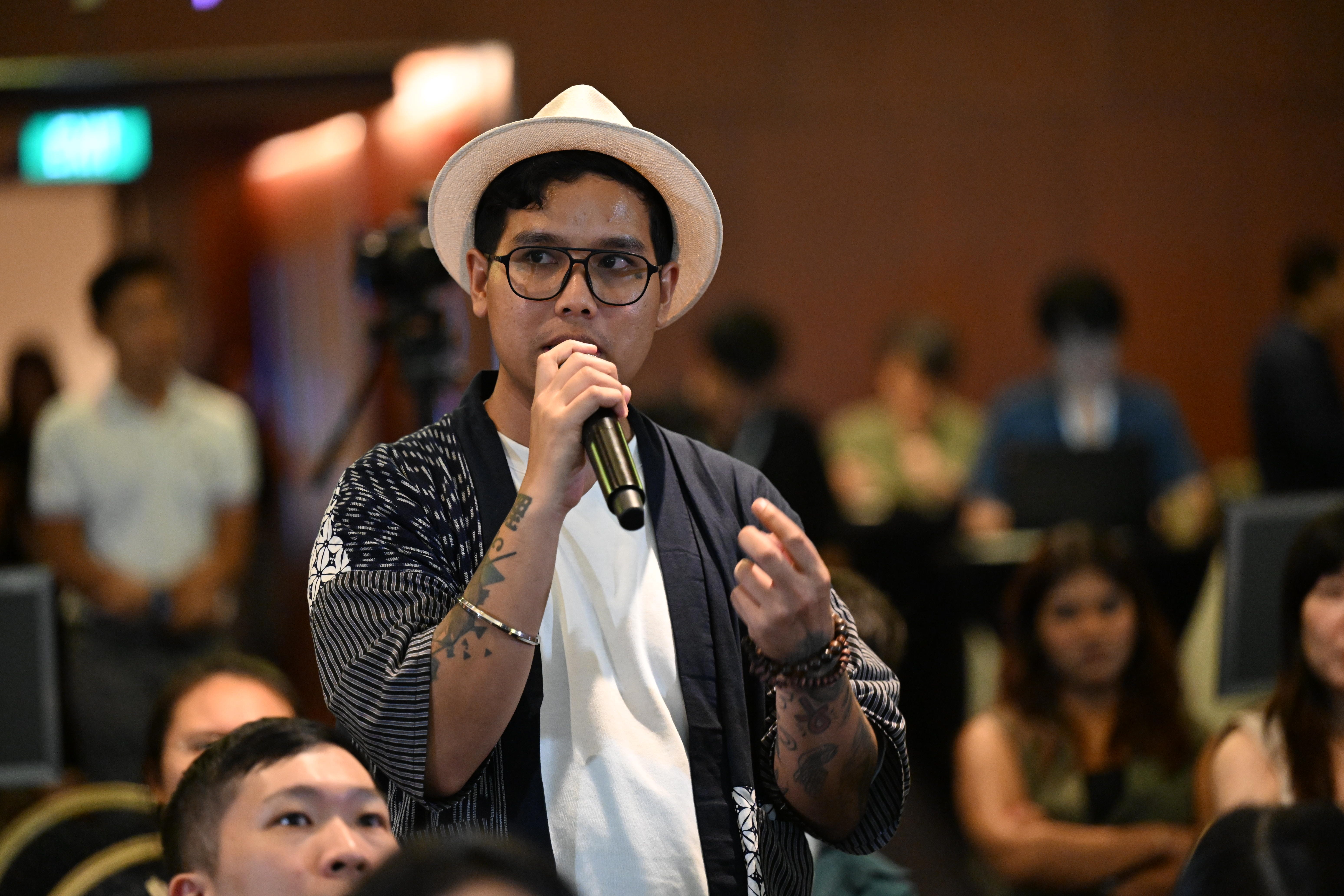 Person with glasses and a hat speaks into a microphone, wearing a patterned jacket. Audience visible.